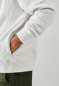 Sudadera blanca texturizada con diseño en relieve, puños acanalados y un bolsillo lateral. Llevada sobre pantalones verdes, mostrando los detalles de la tela.