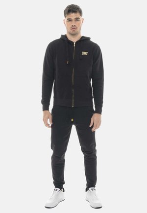 Man die naar voren kijkt, draagt een zwarte hoodie met rits en het "LEONE" logo, bijpassende zwarte sweatpants en witte sneakers op een witte achtergrond.
