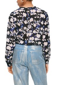 Bloemige blouse met zwarte, witte en blauwe patronen; lange mouwen; keyhole-detail aan de achterkant. Gecombineerd met glanzende lichtblauwe jeans.