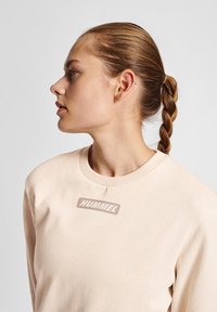 Lys beige sweatshirt med ribbet rund hals, der har et diskret "HUMMEL" logo i lys grå på forsiden. Glat bomuldsstof.