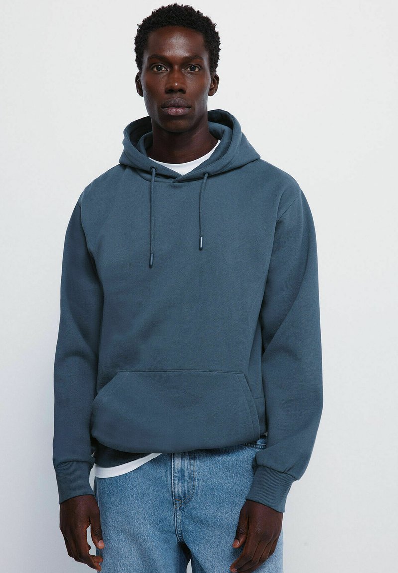 Sweat à capuche bleu doux, avec une poche kangourou, des cordons, des poignets côtelés et une texture lisse, porté par-dessus une chemise blanche.