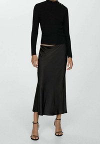 Femme portant un haut noir ajusté à manches longues avec des boutons à l'épaule, une jupe midi en satin noir, et des talons avec bride de cheville noirs, se tenant devant un fond uni.