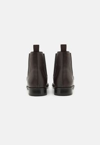 Une paire de bottines en cuir marron foncé avec des languettes, vues de l'arrière sur un fond gris clair uni.