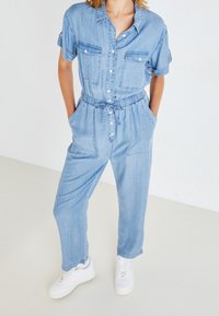 Kvinna i ljus denim jumpsuit med korta ärmar, knäppning fram, dragsko i midjan och vita sneakers, stående med händerna i fickorna.