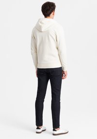 Crème hoodie met een trekkoord capuchon, ribgebreide mouwen en zoom, gecombineerd met donkerblauwe denim jeans en witte sneakers met zwarte accenten.