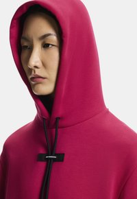 Ružová mikina s kapucňou a šnúrkami, s čiernym ozdobným štítkom. Hladká tkanina s voľným strihom, zakrývajúca časť tváre.