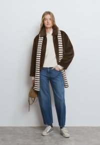 Abrigo marrón de piel sintética con bufanda de rayas en crema y marrón, combinado con un suéter crema, jeans azules y zapatillas blancas con detalles plateados.