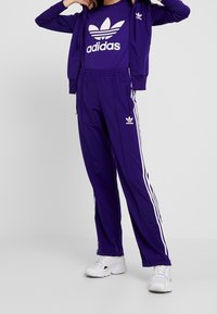 Lila Adidas träningsdräkt med en jacka med dragkedja och byxor med vita ränder, logodetaljer och en bekväm passform. Vita sneakers ingår.