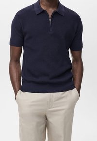 Polo manches courtes bleu marine avec un motif texturé, un col zippé et un ourlet côtelé. Associé à un pantalon beige clair.