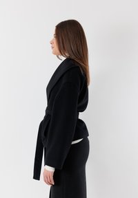 Giacca nera a rappezzo realizzata in tessuto morbido, con ampio colletto e cintura in vita. Indossata sopra un maglione bianco, evidenziando una silhouette strutturata.