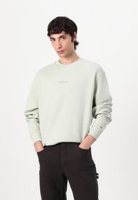 Lys grøn sweatshirt med vaffelmønster, rund hals og broderet logo. Sæt sammen med mørke bukser med synlige lommer.