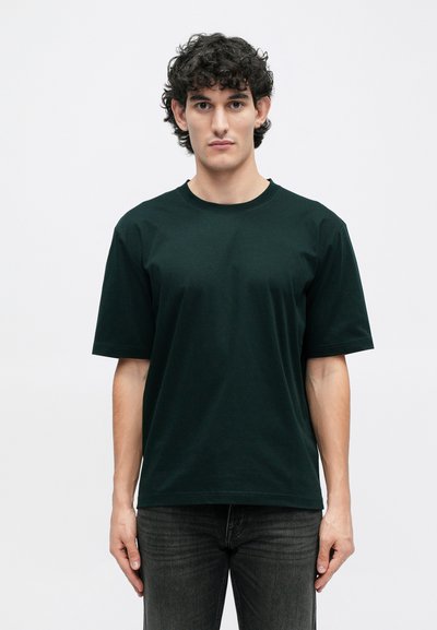 Sötétzöld pamut póló rövid ujjakkal, kerek nyakkivágással, laza szabású, egyszerű anyagtextúrával, sötét farmerral viselve.