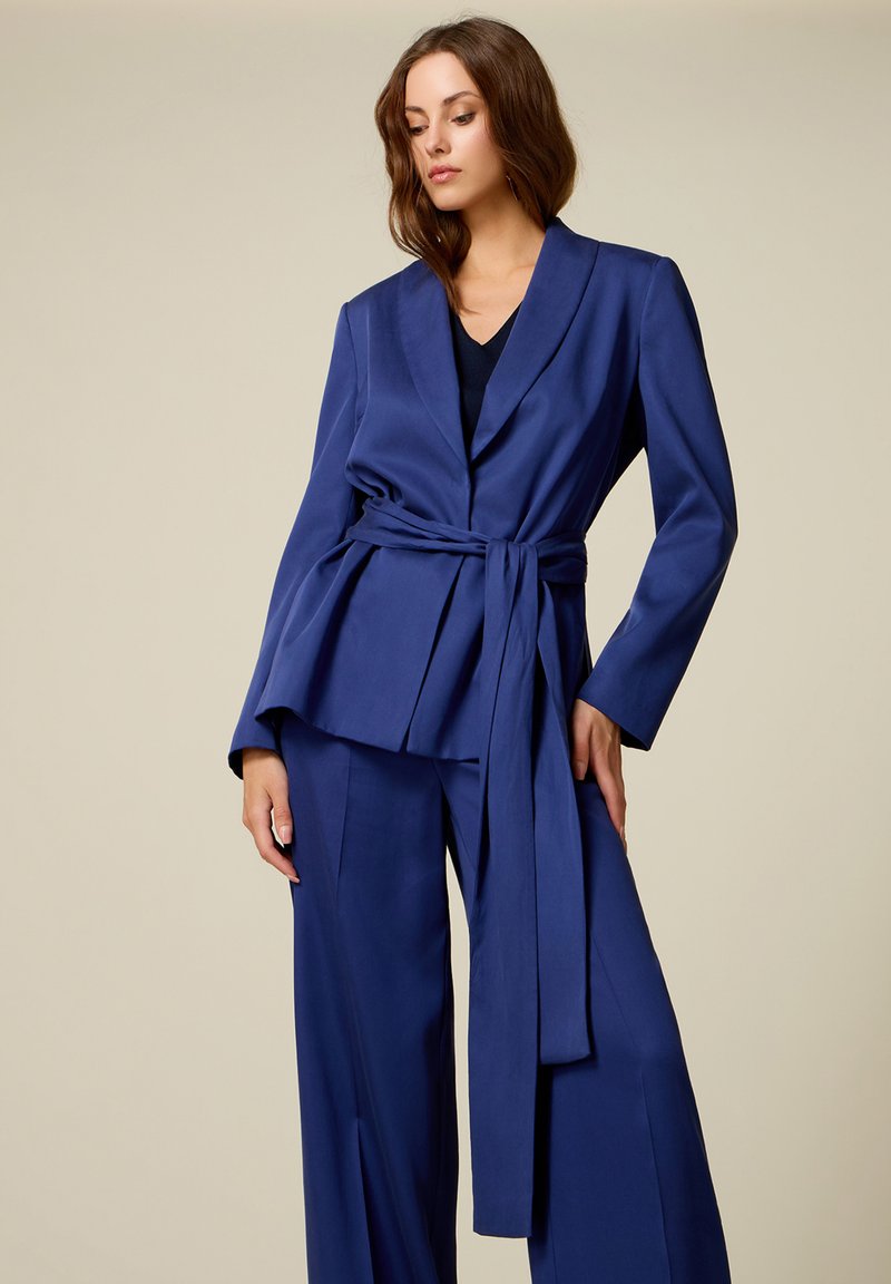 Femme portant un blazer bleu ceinturé et un pantalon large assorti, debout devant un fond beige uni.