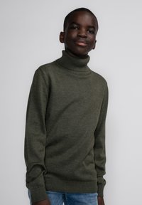Maglione a collo alto verde oliva realizzato in tessuto lavorato a maglia, con maniche lunghe e orlo a coste. Indossato con jeans azzurri chiari, sfondo semplice.