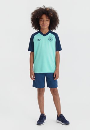 Παιδί με κατσαρά μαλλιά που φοράει αθλητική μπλούζα σε ανοιχτό γαλάζιο και σκούρο μπλε, σκούρα μπλε σορτς και σκούρα μπλε αθλητικά παπούτσια, στέκεται και κοιτάει εμπρός.