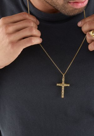 Mann in schwarzem Hemd hält eine goldene Halskette mit einem kunstvoll gestalteten goldenen Kreuzanhänger nahe seiner Brust.