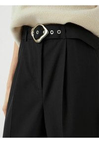 Černé kalhoty na míru mají unikátní stříbrnou sponu na opasek, plisovaný přední díl a jemnou texturu, navržené s kovovými očky pro zapínání.