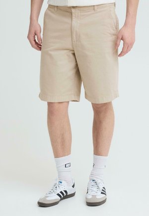 Mann iført beige shorts til knærne, hvite crew-sokker og hvite joggesko med svarte striper, stående på hvit bakgrunn.