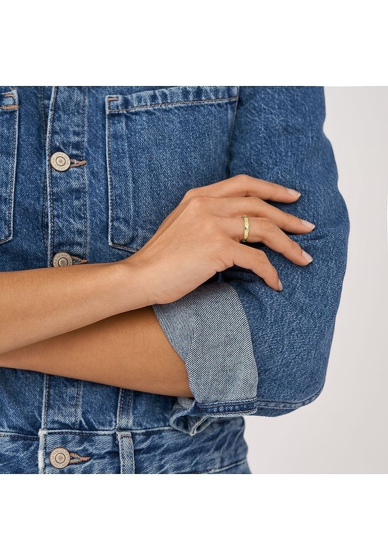 Personne portant une veste en jean bleue avec les manches retroussées, montrant les bras croisés et une bague en or au doigt annulaire de la main droite.