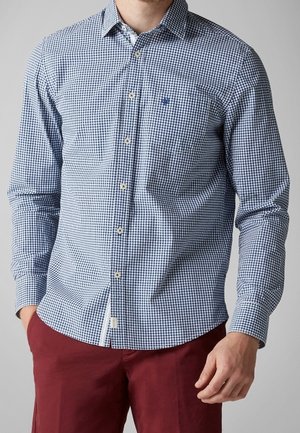Mann trägt ein blau-weiß kariertes langärmliges Hemd mit Knöpfen, kombiniert mit roter Hose, Hände ruhen nahe den Taschen.