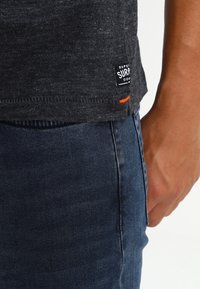 Tmavě šedé tričko s texturovaným povrchem, s malou značkovou nálepkou a oranžovým akcentem u lemování, v kombinaci s tmavými džíny.