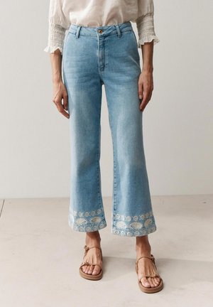 Personne portant un jean court bleu clair avec une broderie florale blanche aux ourlets et des sandales beiges, debout sur un sol clair.