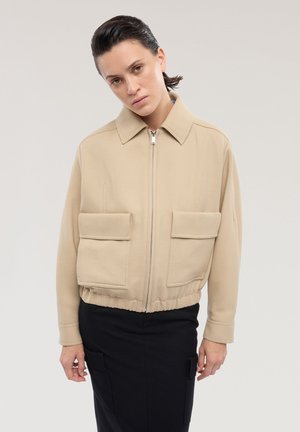 Kvinde med glat tilbageglat mørkt hår iført en beige lynlåstrøje med store forlommer og et sort skørt mod en ensfarvet baggrund.