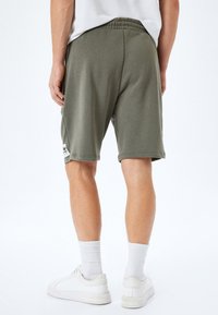 Mand iført olivengrønne shorts til knæene, hvide sokker og hvide sneakers, stående med ryggen mod kameraet på hvid baggrund.