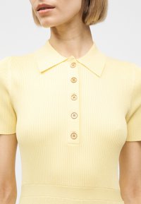 Γυναίκα που φοράει κοντομάνικη μπλούζα σε απαλό κίτρινο με ριγέ υφή, γιακά και πέντε στρογγυλά κουμπιά κατά μήκος της μπροστινής κεντρικής πλευράς.