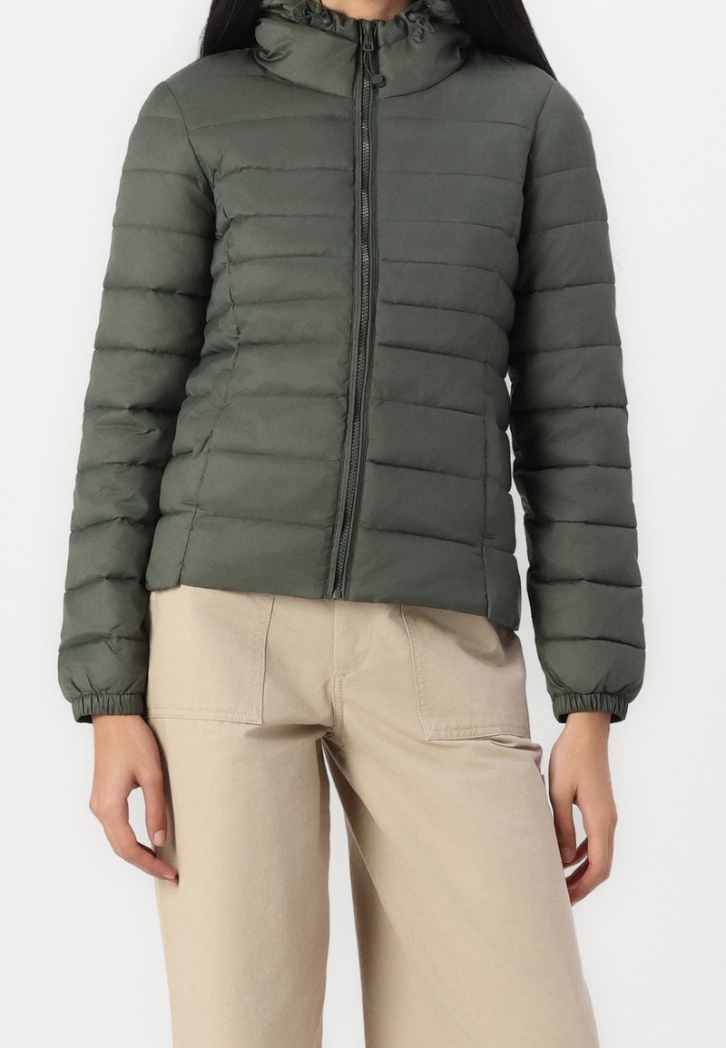 Femme portant une veste matelassée verte foncée zippée avec des poignets élastiques et un pantalon large beige contre un fond uni.