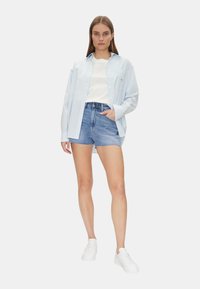 Camicia azzurra a righe con bottoni, t-shirt bianca e pantaloni Corte in jeans. Sneakers bianche. Design casual e comodo. Dettaglio della tasca sulla camicia.