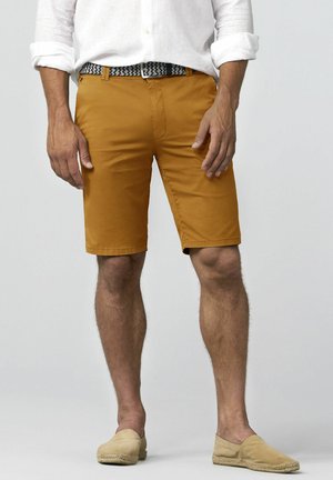 Man met mosterdkleurige shorts, een wit opgerold overhemd, gevlochten riem en beige instapschoenen, staand tegen een effen achtergrond.