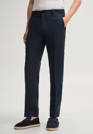 Mężczyzna noszący granatowe spodnie o prostych nogawkach z bocznymi kieszeniami oraz czarne wsuwane mokasyny, stojący na jasnym tle.