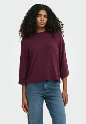 Donna con capelli ricci che indossa una maglia bordeaux a maniche lunghe e jeans blu, in piedi davanti a uno sfondo chiaro uniforme.
