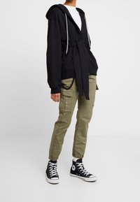 Zwarte hoodie met trekkoorden, ceintuur in de taille, gecombineerd met olijfkleurige cargobroek met meerdere zakken en zwarte hoge sneakers.