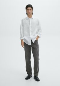 Weißes Leinenhemd mit Knöpfen und hochgekrempelten Ärmeln, kombiniert mit dunkelgrauen Hosen und schwarzen Slippern. Der schlichte Hintergrund betont das Outfit.