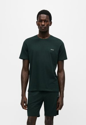 Homme portant un T-shirt à manches courtes vert foncé avec le logo "BOSS" et un short assorti, debout devant un fond clair uni.