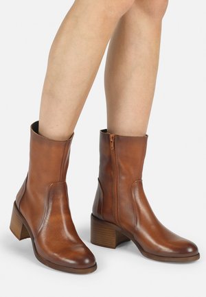 Stivali ankle in pelle marrone con zip laterale, punta arrotondata e tacco in legno impilato. La texture liscia e le linee pulite definiscono il design.