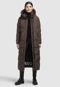 Brūns garš pūšamo mēteli ar kapuci, ar iešūtiem laukumiem, rāvējslēdzēja kabatām un melnu iekšpusi ar baltu zīmolu. Gluda, izturīga materiāla.