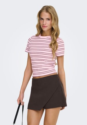 Young woman with long blonde hair wearing a pink striped crop top and dark brown wrap skirt, holding a black handbag strap.
