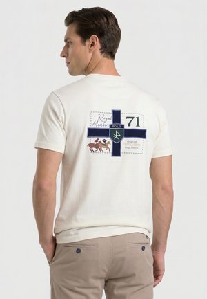 Hombre con pantalones beige y camiseta blanca con cruz azul marino y diseño de jugadores de polo en la espalda, de pie frente a un fondo claro y liso.