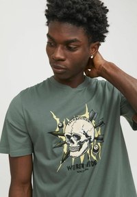 T-shirt en coton vert présentant un grand graphique d'une tête de mort avec des rayons, le texte "JACK & JONES WORLDWIDE" en noir et avec des accents jaunes.