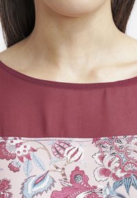 Haut bordeaux à motif floral dans des nuances de rose et de bleu. Texture douce, encolure bateau, et tissu contrastant sur la poitrine.