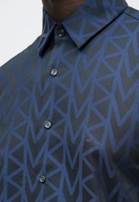 Tmavomodrá košeľa s geometrickým vzorom trojuholníkov v tmavšej modrej farbe, s golierom na gombíky, dvoma gombíkmi a vyšitou modrou značkou "MK".