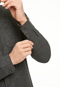 Donkergroene viltshirt met lange mouwen, voorzien van een knoopmanchet en schouderdetail. De stof heeft een zachte textuur en een subtiel patroon.
