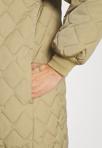 Beżowy pikowany płaszcz z wzorem w romby, z prążkowanymi mankietami i boczną kieszenią, na której spoczywa ręka. Teksturowana tkanina.