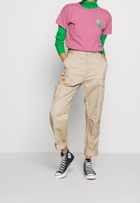 Růžové grafické tričko vrstvené přes zelenou dlouhý rukáv, spárováno s béžovými cargo kalhotami s kapsami a oranžovou nášivkou, a černými teniskami.