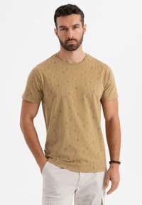 Tan t-shirt med korte ærmer, prydet med et trykt mønster af bogstaver og linjer i mørkere nuancer. Blødt stof med rund halsudskæring.
