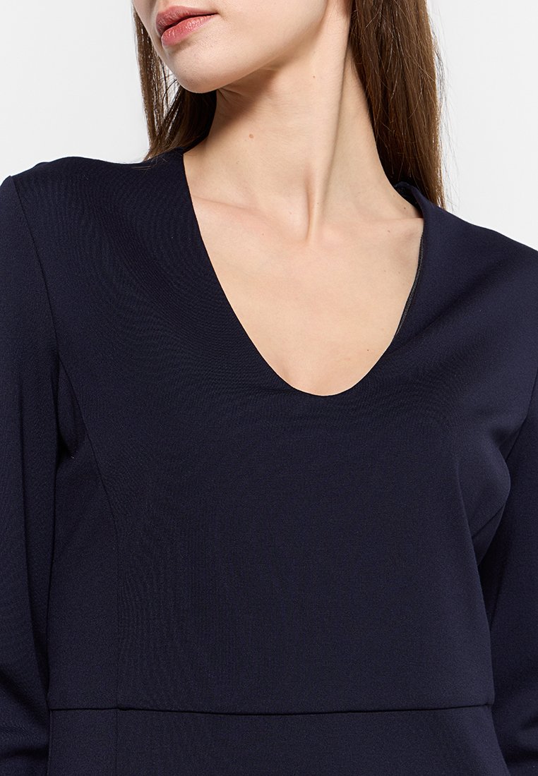 Donna che indossa un top a maniche lunghe V di colore blu navy scuro, mostrata dalle spalle fino a metà torso su uno sfondo bianco.