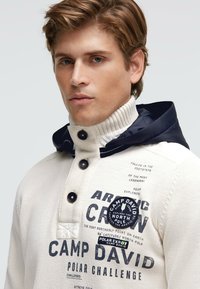 Pullover-Hoodie in Creme mit hohem Kragen, marineblauer Kapuze und Knopfverschluss. Mit gedrucktem Text und Applikationen, strukturiertem Strickmaterial.